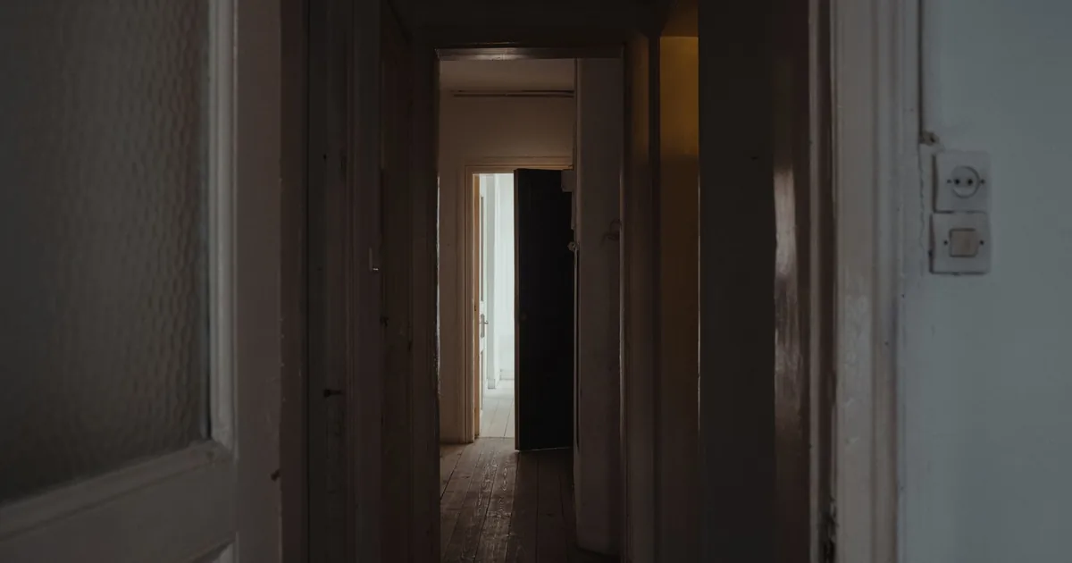 Una porta di legno socchiusa in un corridoio buio, con una lama di luce calda sul pavimento