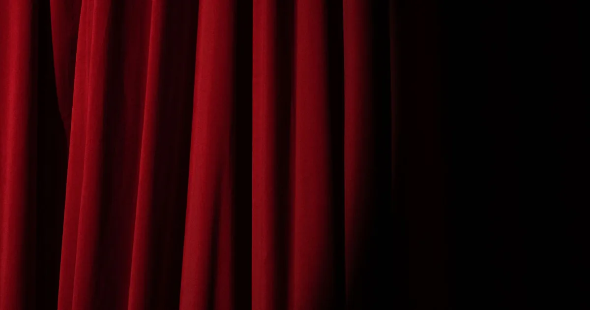 Una sala teatrale vuota e in penombra, con il sipario rosso chiuso e una sola luce sul palco